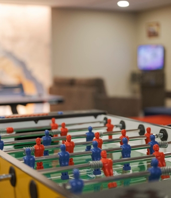 Tischfußball im Familienhotel in Naturns Ein Tischfußballtisch steht in einem Freizeitbereich mit Sitzgelegenheiten und Bildschirmen im Hintergrund.