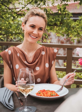 A woman is seated on a terrace, eating pasta with a glass of white wine nearby.