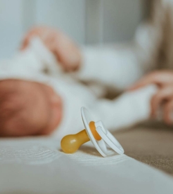 Ein Schnuller liegt auf dem Bett, während im Hintergrund ein Baby unscharf zu sehen ist.