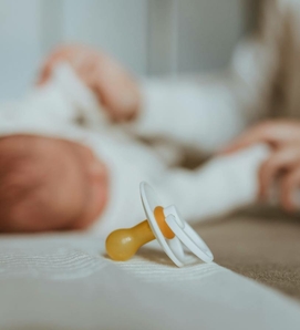 Ein Schnuller liegt auf dem Bett, während im Hintergrund ein Baby unscharf zu sehen ist.