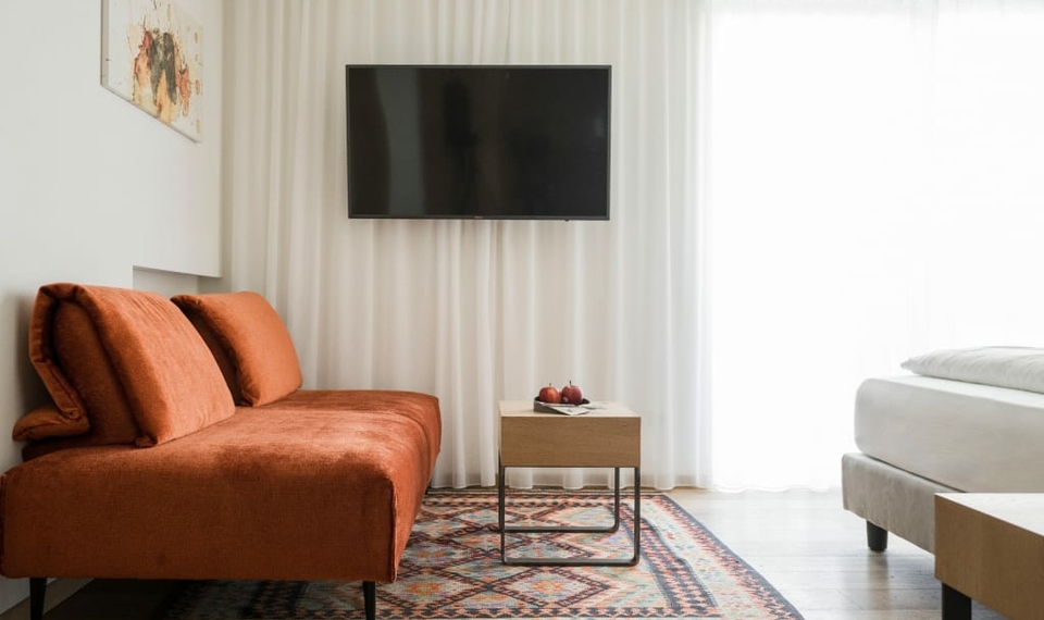 Ein Hotelzimmer mit orangefarbenem Sofa, Wandfernseher, kleinem Tisch und Bett am Fenster.
