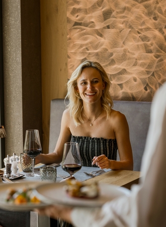 Eine Frau sitzt im Hotelrestaurant und lächelt am gedeckten Tisch, während ein Kellner serviert.