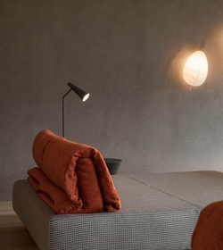 Eine gepolsterte Liege mit orangefarbenen Handtüchern steht in einem Ruheraum neben einer schwarzen Leselampe und einer runden Wandlampe.