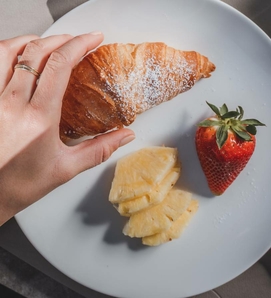Eine Hand hält ein Croissant auf einem Teller mit Ananasstücken und einer Erdbeere.