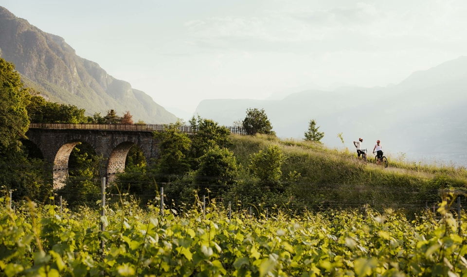 Two mountain bikers ride along a path beside an old stone viaduct in the landscape near Naturns.
