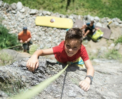 Ein Junge klettert an einer Felswand mit Sicherungsseil, während darunter weitere Personen mit Kletterausrüstung sitzen.