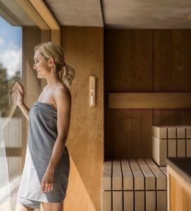 A woman stands in a bright sauna area beside the glass front and looks outside.