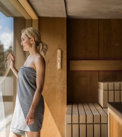 A woman stands in a bright sauna area beside the glass front and looks outside.