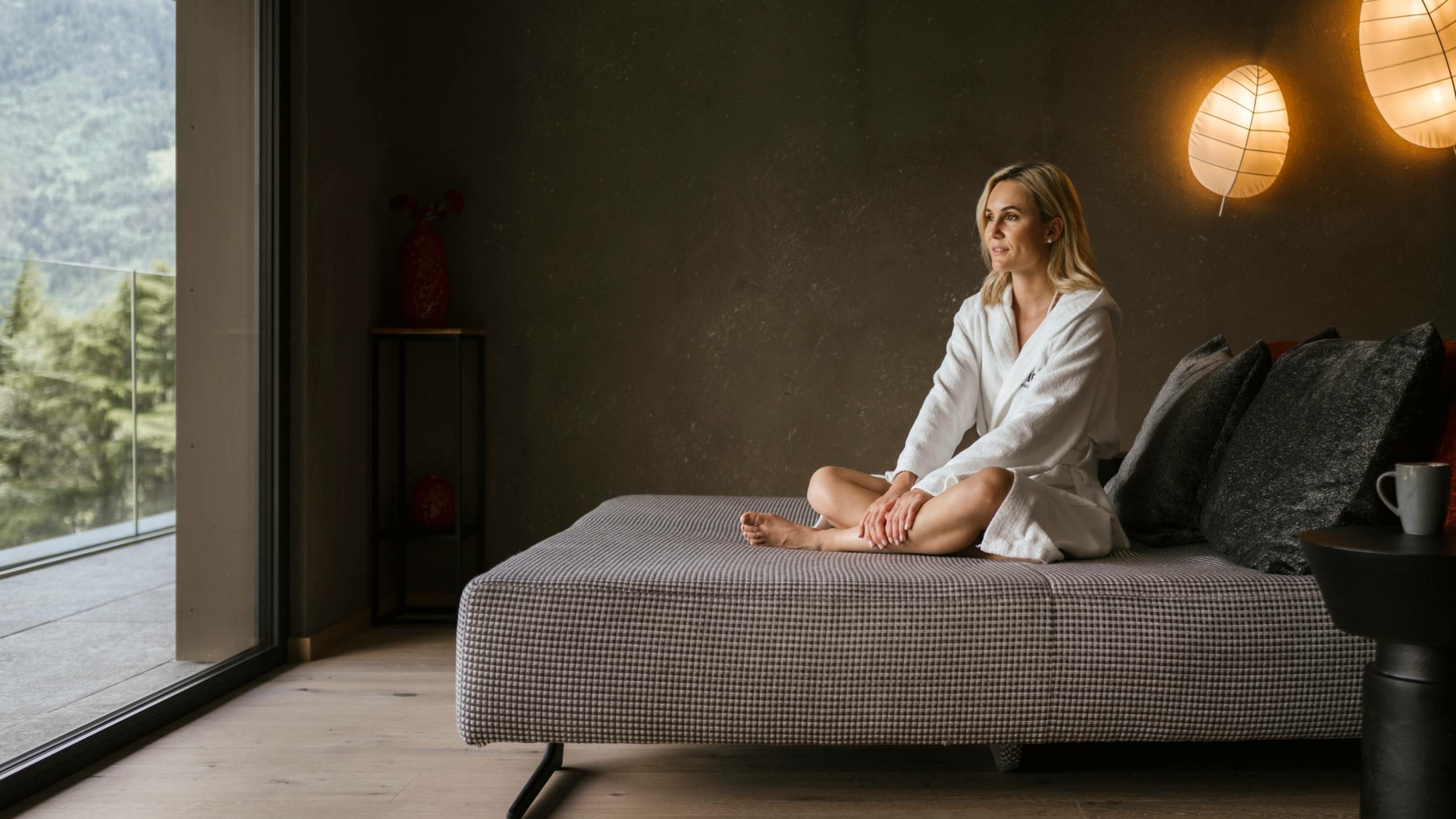 Una donna è seduta su una chaise longue nella sala relax accanto alle grandi finestre con vista sul verde.