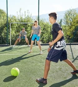 Drei Jugendliche spielen auf einem Kunstrasenplatz Fußball vor einem Tor im Freien.