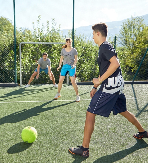 Drei Jugendliche spielen auf einem Kunstrasenplatz Fußball vor einem Tor im Freien.