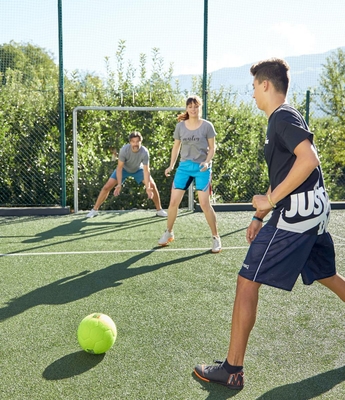 Familienurlaub mit Fußball auf dem Platz Drei Jugendliche spielen auf einem Kunstrasenplatz Fußball, ein Tor und ein grüner Ball sind zu sehen.