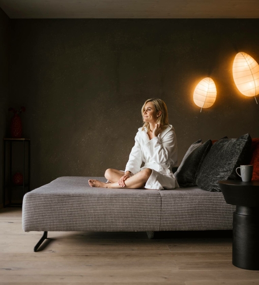 A woman sits on a lounger in the relaxation area of a softly lit wellness room.