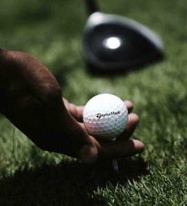 A hand holds a golf ball on a tee against the green turf of a golf course area.