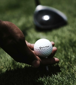 A hand holds a golf ball on a tee against the green turf of a golf course area.