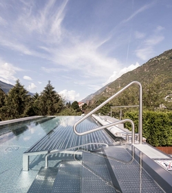 Una piscina infinity con scala in acciaio si trova davanti a un paesaggio montano e a un cielo velato.