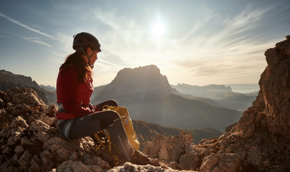 Eine Kletterin sitzt auf einem Felsen und blickt bei Sonnenaufgang über die Berglandschaft.