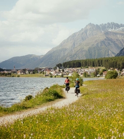 Two cyclists ride along a lakeside path, with mountains and the village in the background.