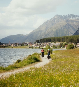 Two cyclists ride along a lakeside path, with mountains and the village in the background.