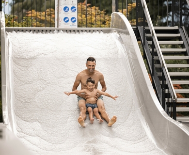 Rutschbahn im Wellnesshotel Feldhof in Naturns Ein Mann und ein Kind rutschen gemeinsam auf einer weißen Wasserrutsche im Hotelbereich.