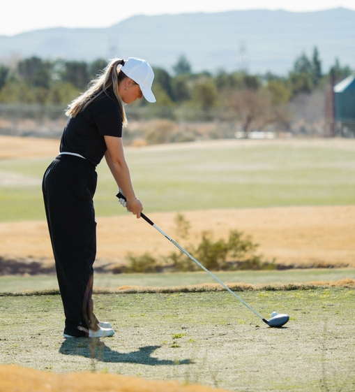 A female golfer lines up her shot on the tee box at a golf course with hills in the background.