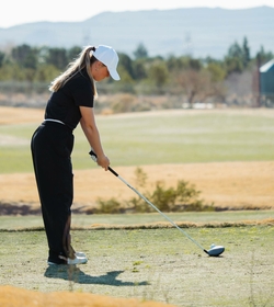 A female golfer lines up her shot on the tee box at a golf course with hills in the background.