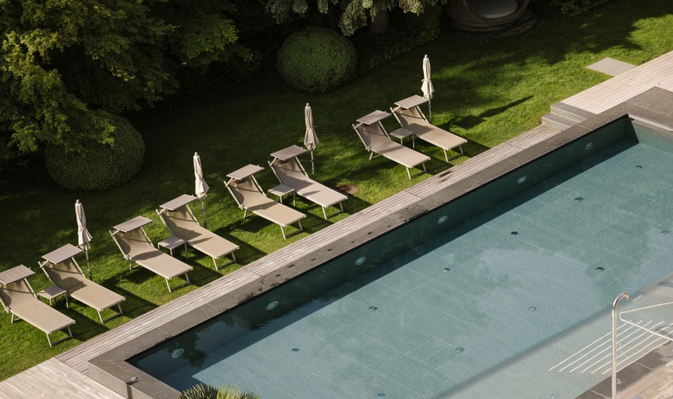 Una piscina all'aperto con diversi lettini e ombrelloni lungo il bordo del prato, davanti a una vegetazione fitta.