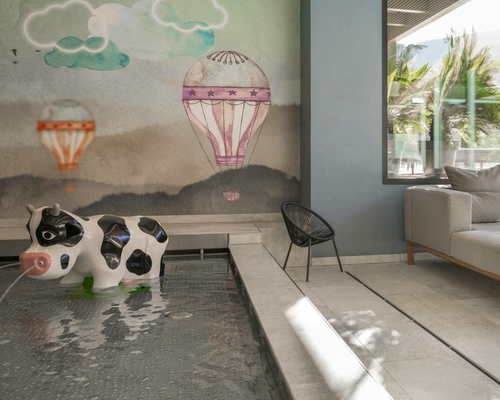 Ein kleiner Pool mit Kuh-Sitzfigur steht vor einer Wandmalerei mit Heißluftballons und Wolken.