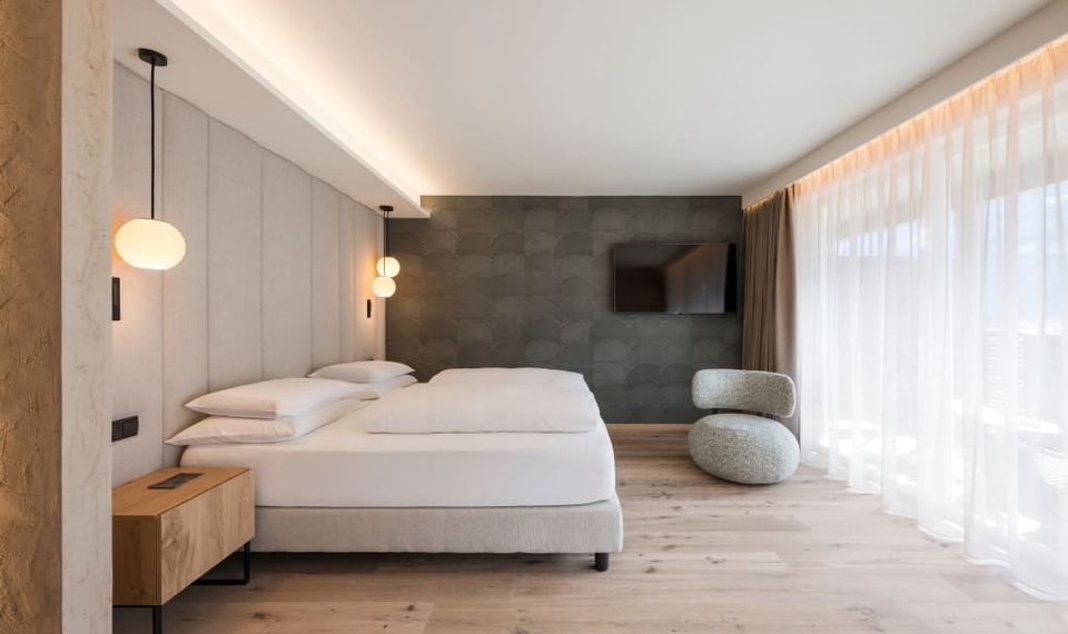 Suite Harmonie im Feldhof: modernes Schlafzimmer Ein modernes Schlafzimmer mit Doppelbett, hellen Vorhängen, Wandlampen und einem Sitzhocker am Fenster.