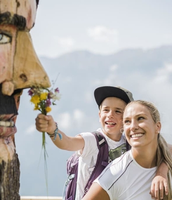 Vacanza escursionistica con maschera di legno Un bambino porge un piccolo mazzo di fiori a una donna sorridente durante un'escursione in montagna, accanto a una maschera di legno intagliata.