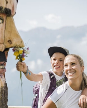 Ein Kind hält einer lächelnden Frau auf einer Bergwanderung einen kleinen Blumenstrauß hin, neben einer geschnitzten Holzmaske.