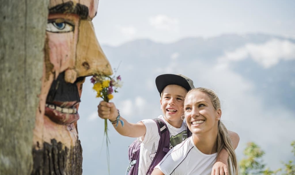 Wanderurlaub mit geschnitzter Holzmaske Ein Kind hält einer lächelnden Frau auf einer Bergwanderung einen kleinen Blumenstrauß hin, neben einer geschnitzten Holzmaske.
