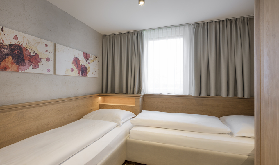 Suite mit Doppelbett und Einzelbett im Feldhof Ein schlichtes Hotelzimmer mit einem Doppelbett, einem Einzelbett, hellem Fenster und Vorhängen.