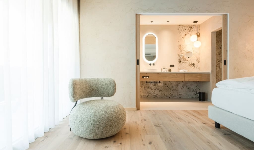 Ein helles Doppelzimmer mit Blick in das offene Bad und einem gepolsterten Sessel am Fenster.