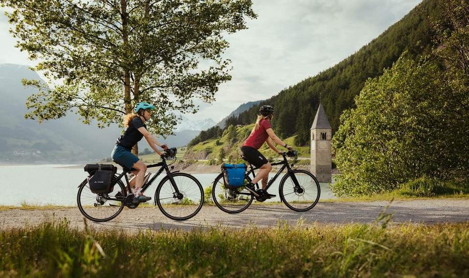 Two people ride e-bikes along a lakeside path, with mountains and a small tower in the background.
