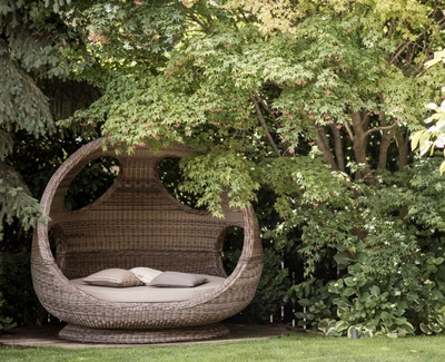 A woven relaxation cocoon with cushions stands in a green garden beneath dense foliage.