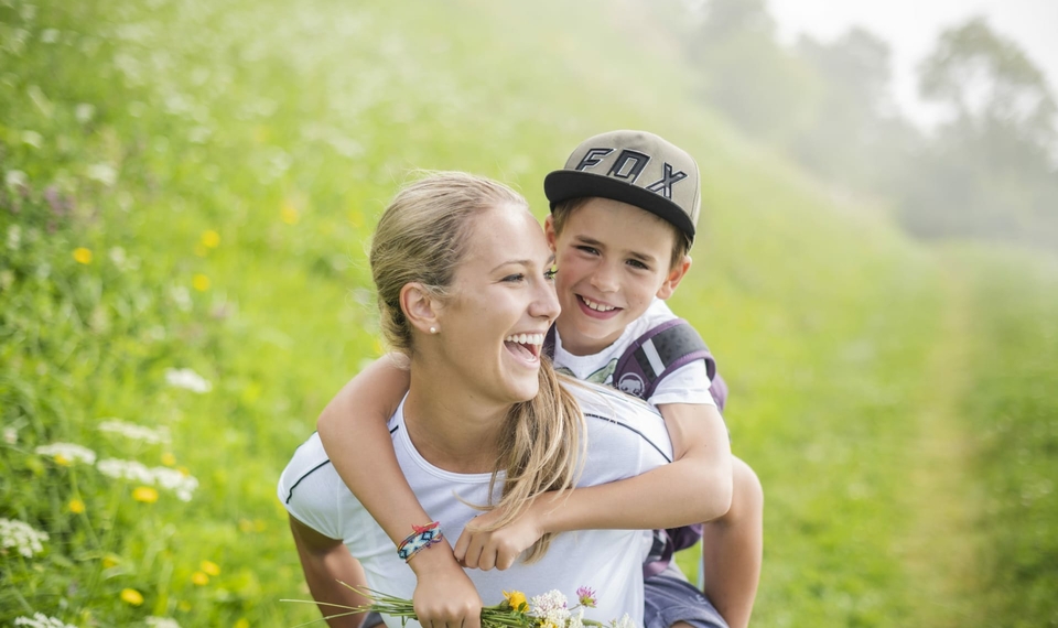 Vacanza in famiglia con escursioni in Alto Adige Una madre porta un bambino sorridente sulla schiena lungo un sentiero verde.