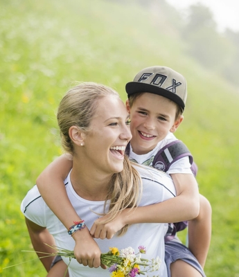 Vacanza in famiglia con escursioni in Alto Adige Una madre porta un bambino sorridente sulla schiena lungo un sentiero verde.
