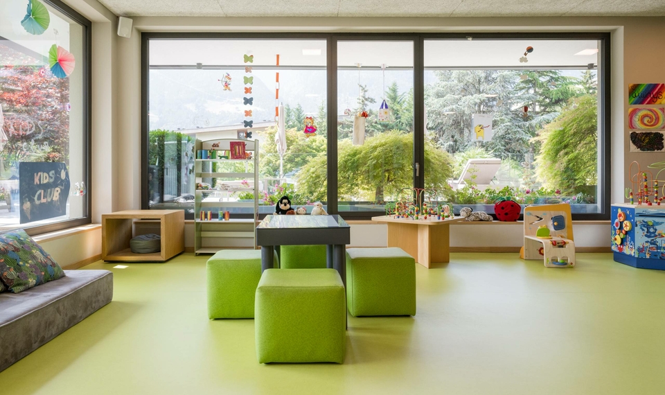 Ein heller Spielraum mit grünen Hockern, Spieltischen und Blick in den Garten für Kinderbetreuung im Hotel.