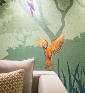 Ein großes Papageien-Wandbild über einer hellen Sitzbank mit Kissen in einem ruhigen Innenraum.