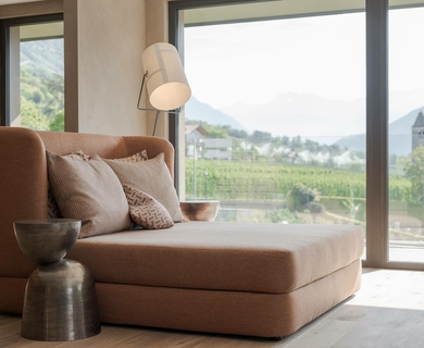 Suite mit Panoramablick im Feldhof DolceVita Resort Eine helle Suite mit Tagesbett, Stehleuchte und großem Fenster mit Blick ins Grüne und auf einen Kirchturm.