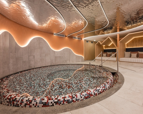 Una sala relax della spa con piscina rotonda a mosaico, lettini e rivestimento in legno illuminato.