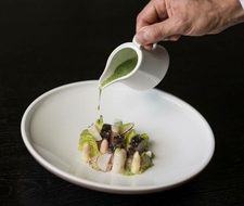 A hand pours green sauce over an elegant plated gourmet dish on a white plate.