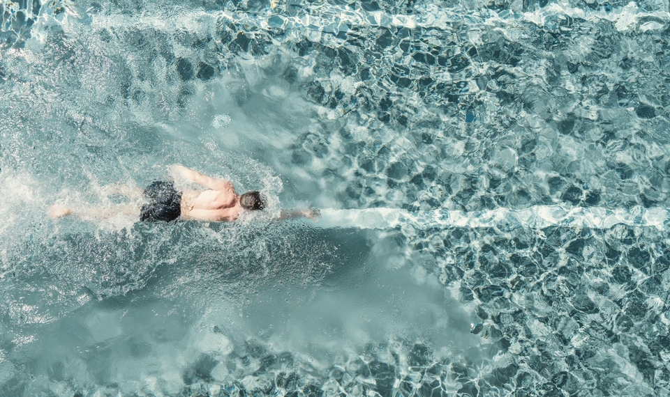 Una persona nuota nell'acqua limpida della piscina con riflessi di luce e fondo piastrellato visibile.