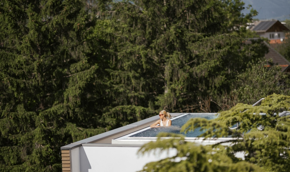 Eine Person schwimmt im Sky Infinity Pool auf dem Dach, umgeben von Bäumen und Blick auf Häuser im Hintergrund.