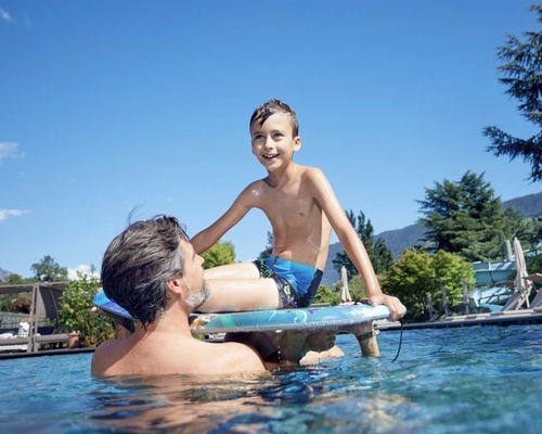 Ein Junge sitzt auf einem Schwimmboard im Pool, während ein Mann ihn im Wasser stützt.