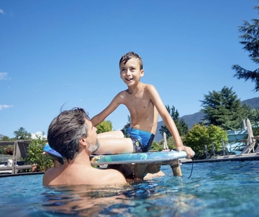 Ein Junge sitzt auf einem Schwimmboard im Pool, während ein Mann ihn im Wasser stützt.