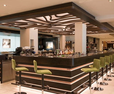 Hotelbar im Feldhof DolceVita Resort Eine moderne Hotelbar mit grün gepolsterten Barhockern und beleuchtetem Tresen im Feldhof DolceVita Resort.