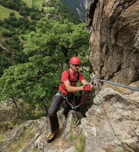 A man in red gear is clipped into a cable while climbing a rock face on a via ferrata.