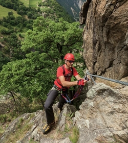 A man in red gear is clipped into a cable while climbing a rock face on a via ferrata.